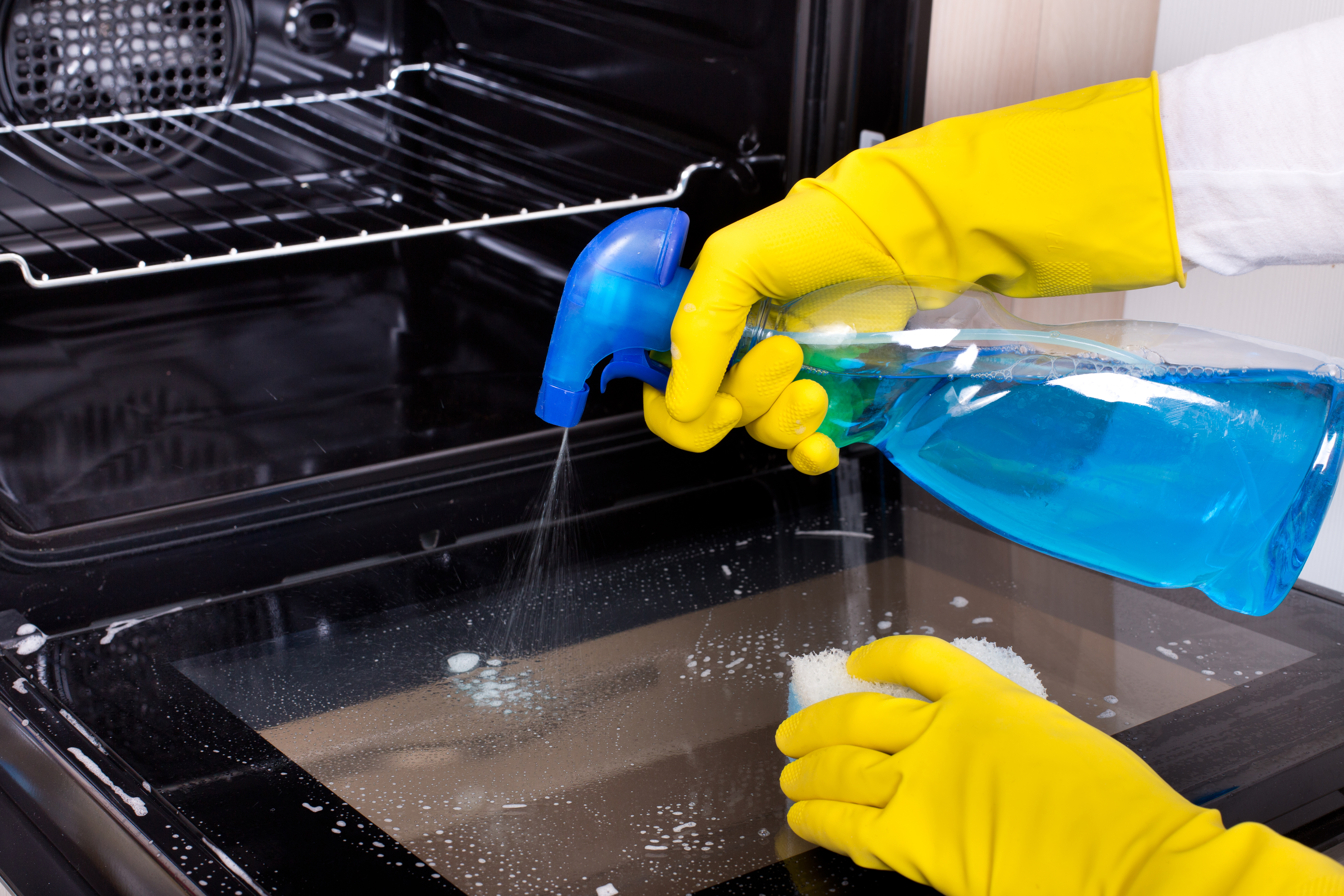 Close up of hand with yellow protective gloves spraying detergent from bottle on the oven door